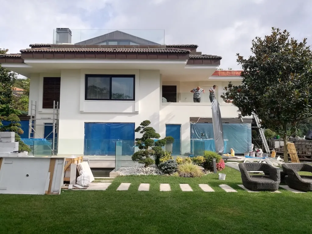 Koruyucu örtüyle kapatılmış bir villa cephesinde çalışan ustalar; kapsamlı yenileme işi sürerken cephe brandayla korunuyor